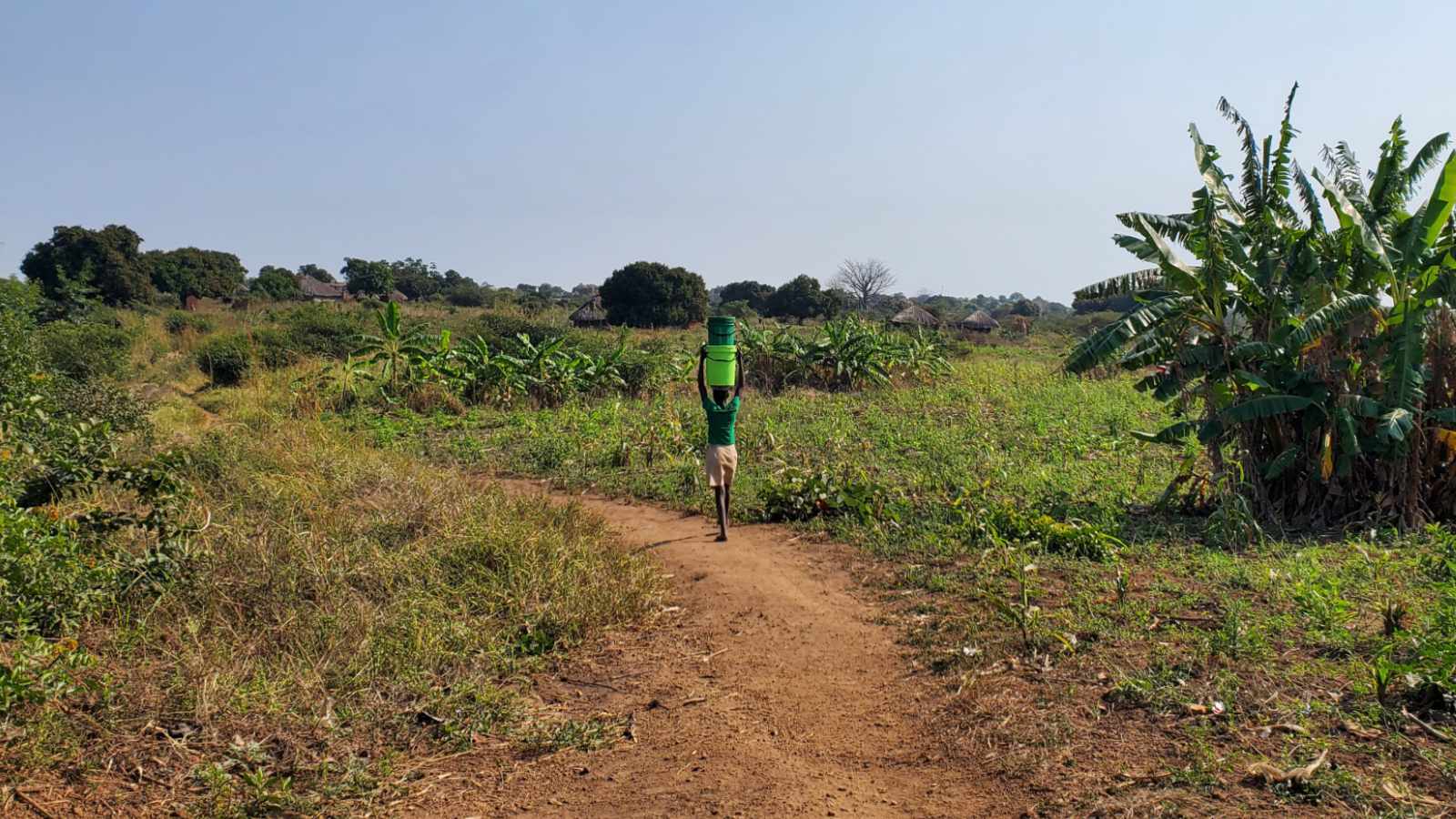 A young girl carries water on her head along a footpath through farmland toward a rural Mozambican village