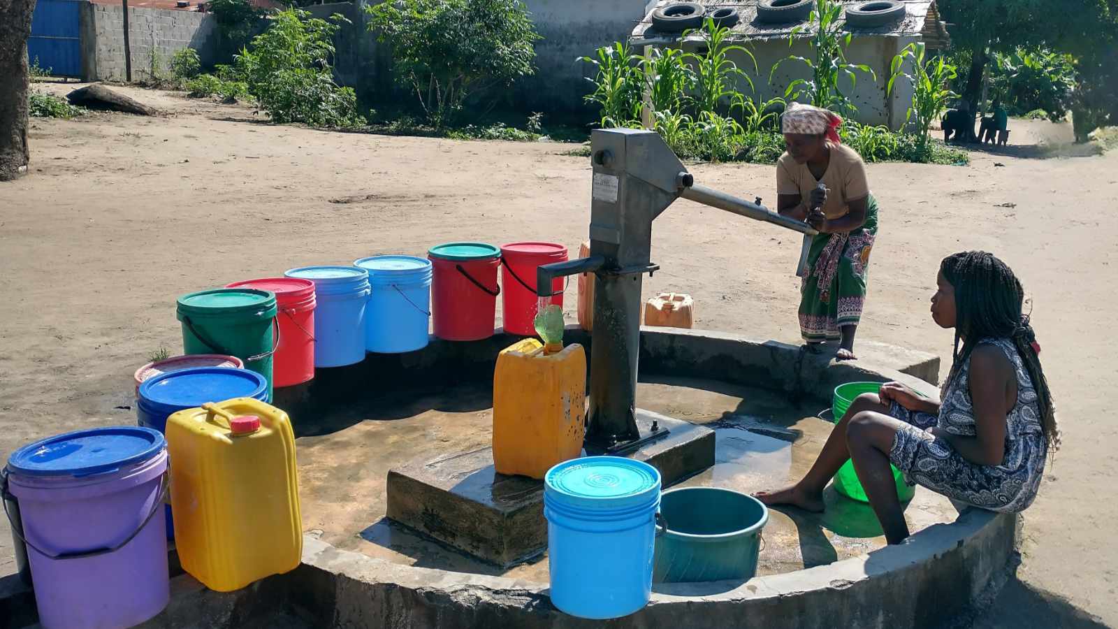 Women drawing water from a hand-pump well surrounded by brightly colored buckets in a Mozambican community