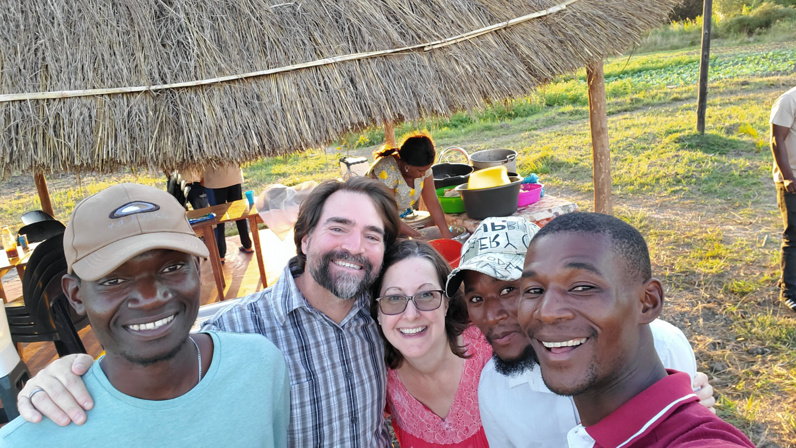 Jason and Susan with friends and partners gathered for a meal under a thatch shelter beside the field in Mozambique