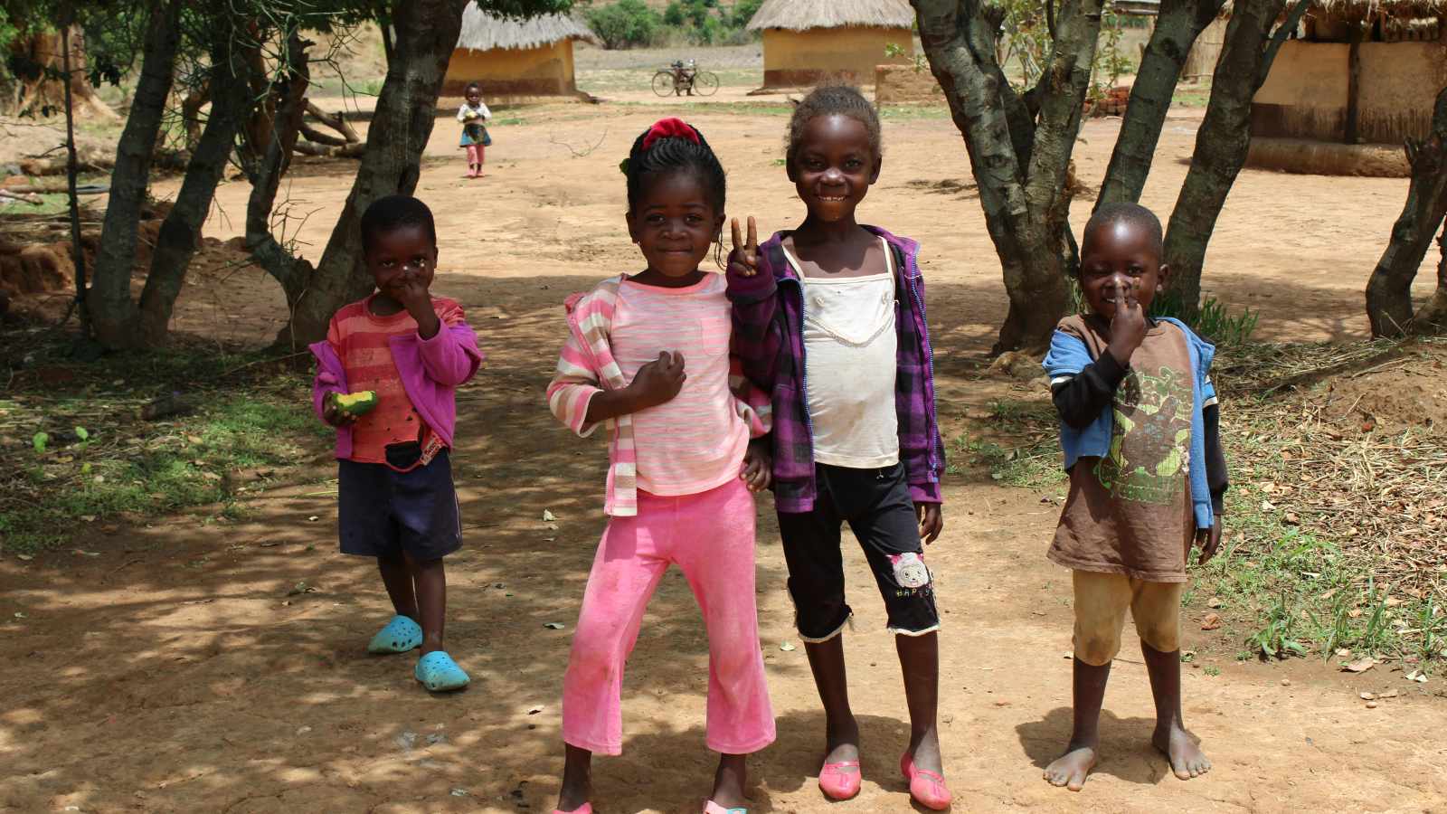 Four smiling children standing on a dirt path in a rural Zambian village