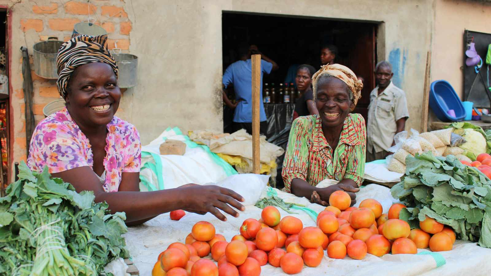 Two women laughing behind a market stall of tomatoes and vegetables in Zambia
